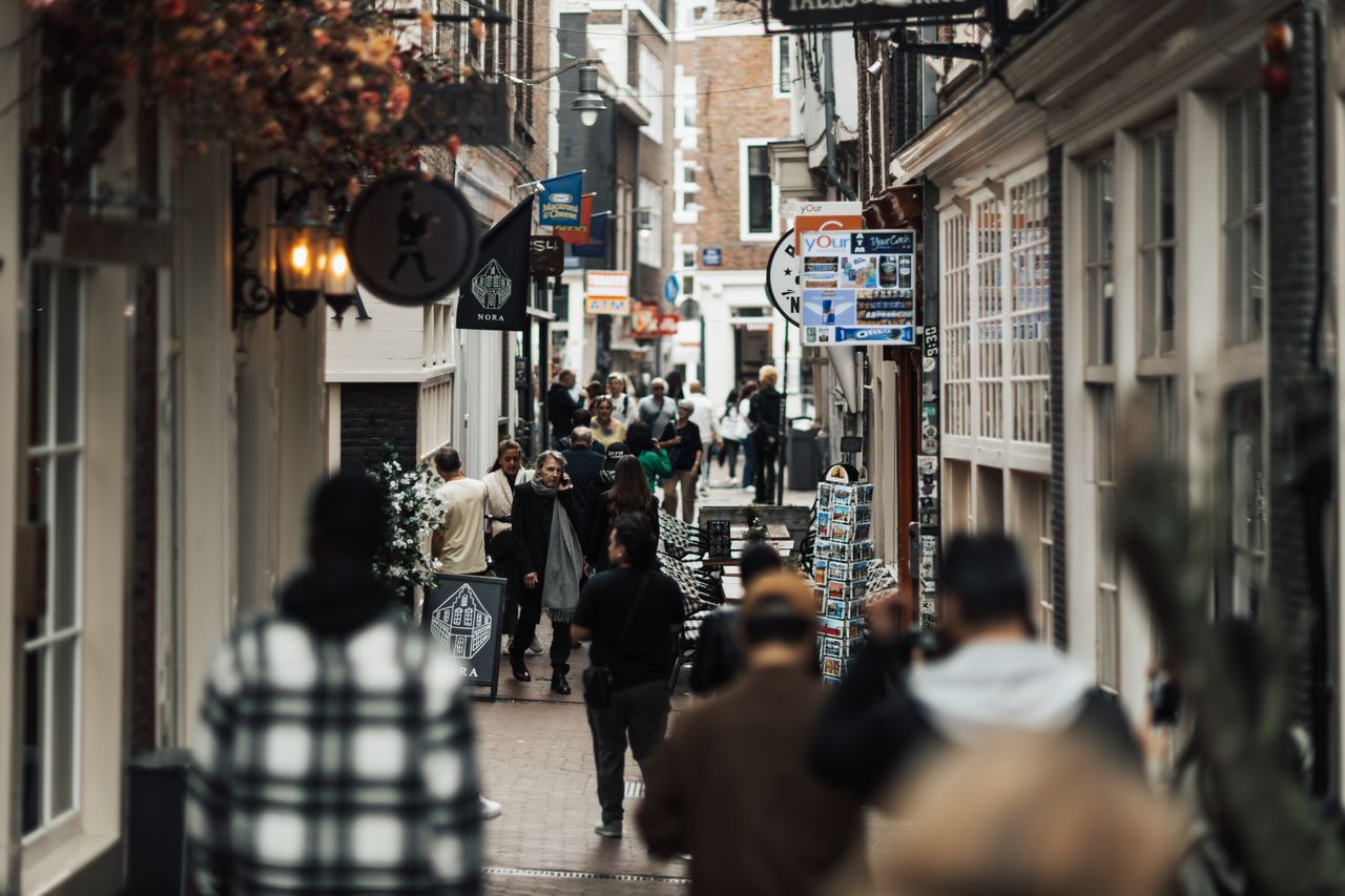 home-hero People walking through a narrow urban alley with shops and signs on both sides.